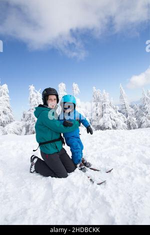 Fröhliche Familie, Mama und Kind in warmen Kleidern, die Spaß im Freien haben und an einem sonnigen, kalten Tag in den Bergen Skifahren lernen auf einer verschneiten Piste. Genießen Sie Wint Stockfoto