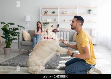 Glücklicher junger arabischer Kerl, der mit Hund spielt, Spaß auf dem Boden zu Hause hat, seine Freundin trinkt Kaffee auf dem Hintergrund Stockfoto