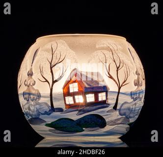 Eine handbemalte Glaskanne mit Winterlandschaft, die von einer Kerze im Inneren der Tasse angezündet wird. Eine Nahaufnahme und die Verwendung von Lichtmalerei. Stockfoto