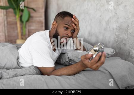 Schockierter schwarzer Mann, der die Uhr im Bett ansah Stockfoto