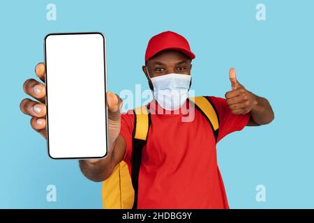 Sichere Lebensmittelzustellung. Schwarzer Kurier Mann Trägt Medizinische Maske Zeigt Blank Smartphone Stockfoto