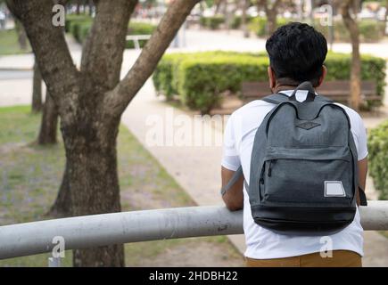 Rückaufnahme eines männlichen Studenten, der sich mit einem Rucksack auf ein Geländer lehnte und in einen Park schaute Stockfoto