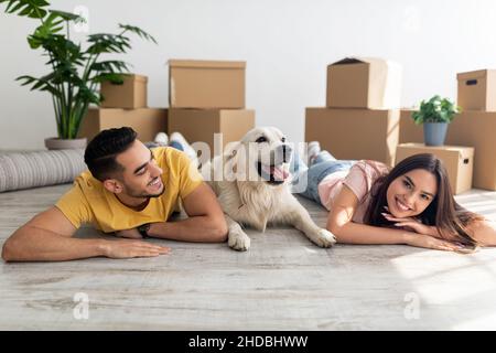 Cooles junges, vielfältiges Paar, das mit seinem niedlichen Hund auf dem Boden zwischen Kartons liegt und mit seinem Haustier in ein neues Zuhause zieht Stockfoto