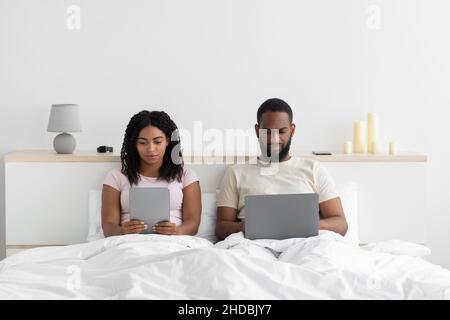 Beschäftigt junge schwarze Frau mit Tablet ignoriert Mann mit Computer, sitzen auf dem Bett im Schlafzimmer Innenraum am Abend Stockfoto