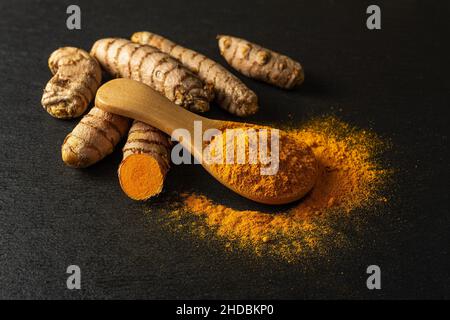 Organisches Kurkuma-Pulver in einem Holzlöffel und rohe Curcuma-Wurzeln auf schwarzem Schiefergrund. Frische und gemahlene Rhizome von Curcuma longa linn. Indische s Stockfoto