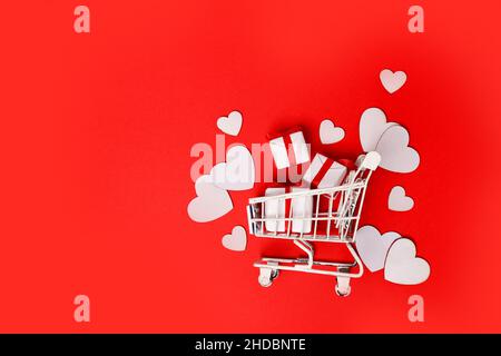 Shopping Trolley mit Geschenkboxen, Liebesherzen auf rotem Hintergrund. Shopping und Verkauf am Valentinstag Stockfoto