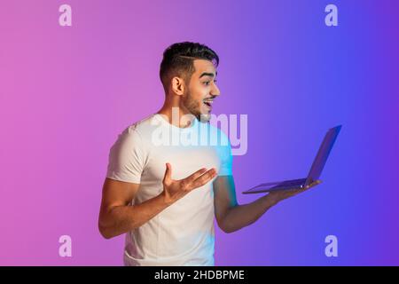 Hübscher junger arabischer Kerl, der auf den Laptop-Bildschirm schaut, begeistert über Online-Lotteriegewinn oder Erfolg im Neonlicht Stockfoto