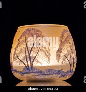 Eine handbemalte Glaskanne mit Winterlandschaft, die von einer Kerze im Inneren der Tasse angezündet wird. Eine Nahaufnahme und die Verwendung von Lichtmalerei. Stockfoto