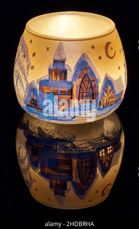 Eine handbemalte Glaskanne mit Winterlandschaft, die von einer Kerze im Inneren der Tasse angezündet wird. Eine Nahaufnahme und die Verwendung von Lichtmalerei. Stockfoto