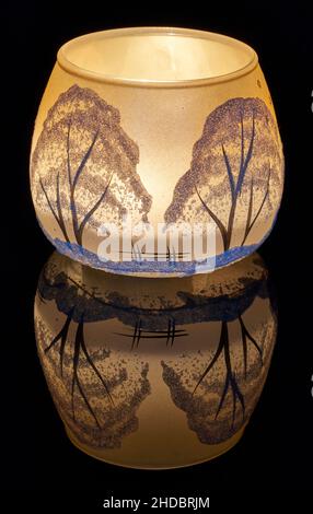 Eine handbemalte Glaskanne mit Winterlandschaft, die von einer Kerze im Inneren der Tasse angezündet wird. Eine Nahaufnahme und die Verwendung von Lichtmalerei. Stockfoto