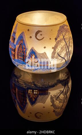 Eine handbemalte Glaskanne mit Winterlandschaft, die von einer Kerze im Inneren der Tasse angezündet wird. Eine Nahaufnahme und die Verwendung von Lichtmalerei. Stockfoto