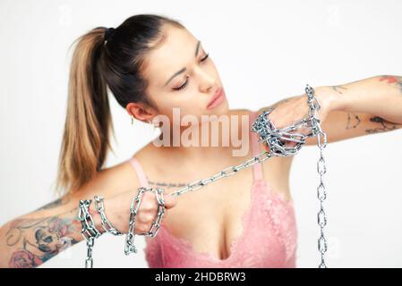 Tätowierte Frau im BH, die eine Stahlkette auf weißem Hintergrund hält und reißt. Mädchenpower-Konzept. Stockfoto