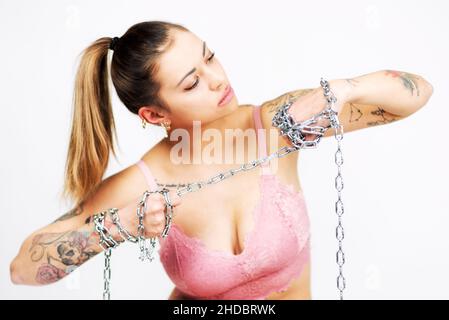 Tätowierte Frau im BH, die eine Stahlkette auf weißem Hintergrund hält und reißt. Mädchenpower-Konzept. Stockfoto