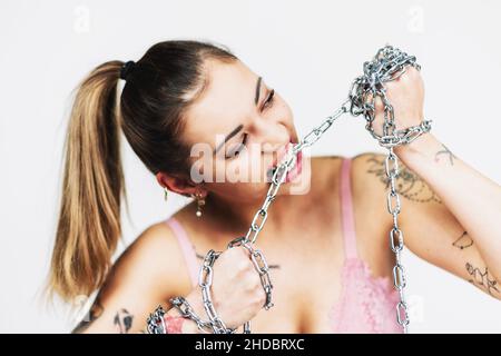 Tätowierte feministische Frau im BH beißt und reißt eine eiserne Kette auf weißem Hintergrund. Mädchenpower-Konzept. Stockfoto