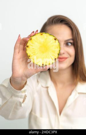 Eine junge, attraktive, hübsche, kaukasisch lächelnde Frau hält eine Ringschneide-Ananas, die ihr Auge vor einem weißen Hintergrund bedeckt. Gesunde Ernährung Konzept Stockfoto