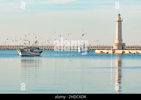 Alte Fischerboote, die vom Fischfang zurückkehren, kommen bei Sonnenuntergang in den Hafen, begleitet von einer Schar von Möwen. Mit schönem Leuchtturm im Hintergrund Stockfoto