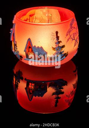 Eine handbemalte Glaskanne mit Winterlandschaft, die von einer Kerze im Inneren der Tasse angezündet wird. Eine Nahaufnahme und die Verwendung von Lichtmalerei. Stockfoto