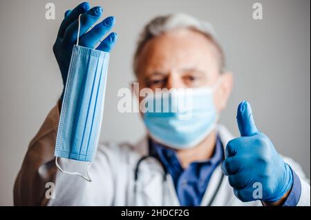 Arzt hält eine Gesichtsmaske und zeigt die Daumen nach oben Stockfoto