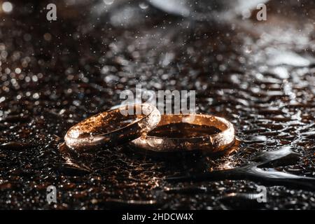 Zwei goldene Eheringe in Regentropfen auf schwarzem, nassem Hintergrund. Stockfoto