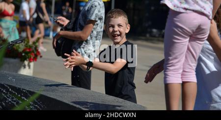 Kinder am Brunnen. Ein achtjähriger Junge macht sich zwischen den Wasserbächen im Springbrunnen lustig. Sommer in der Stadt an einem heißen Tag. Stockfoto