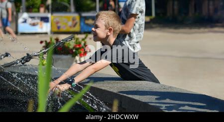 Kinder am Brunnen. Ein achtjähriger Junge macht sich zwischen den Wasserbächen im Springbrunnen lustig. Sommer in der Stadt an einem heißen Tag. Stockfoto