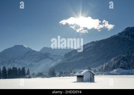 Winteraufnahme einer Scheune im verschneiten Feld, Berge im Hintergrund und Sonnenstrahlen hinter einer Wolke Stockfoto