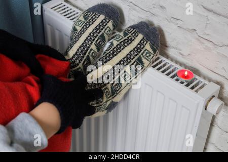 Der Besitzer in warmen Socken und Fäustlingen wärmt sich am Heizkörper auf. Harter Winter. Heizungskrise Stockfoto