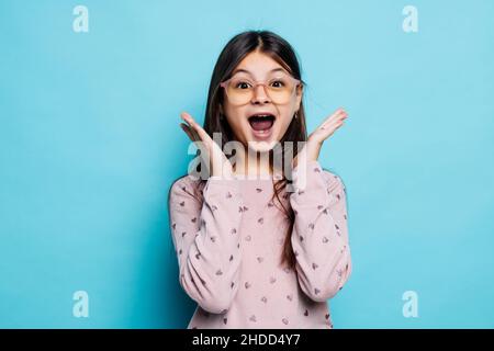 Porträt eines verzweifelten und schockierten schönen kleinen Mädchens mit blauem T-Shirt auf blauem Hintergrund, das die Hände in der Nähe des Gesichts hält, mit weit geöffnetem Mund. Stockfoto