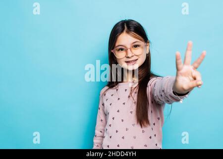 Schöne kaukasische kleine Mädchen trägt gestreiftes Kleid auf blauem Hintergrund zeigt und zeigt mit den Fingern Nummer drei, während lächelnd zuversichtlich ein Stockfoto