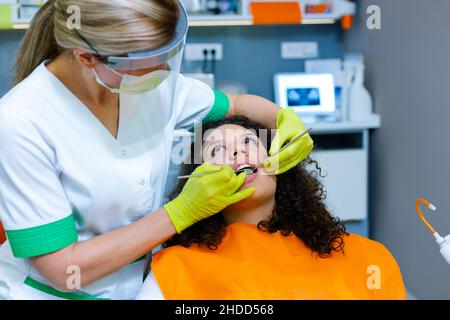 Schönes gemischtes Teenager-Mädchen auf zahnärztliche Untersuchung durch kaukasische Frau mittleren Alters mit Gesichtsvisier und Maske als Coronavirus-Sicherheitsmaßnahme. Stockfoto