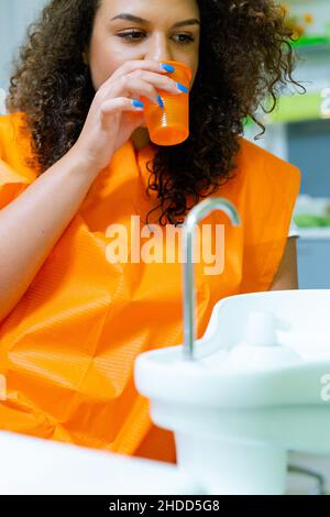 Teenager-Mädchen spülen Mund in der Zahnarztpraxis, Trinkwasser aus Kunststoff orange Farbbecher. Stockfoto