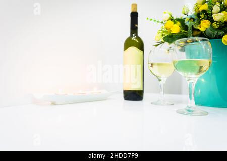 Weißweinflasche mit Balnk-Etikett, zwei gefüllte Gläser und brennende Kerzen auf weißem Tisch mit weißem Wandhintergrund. Romantische Hochzeit Stockfoto