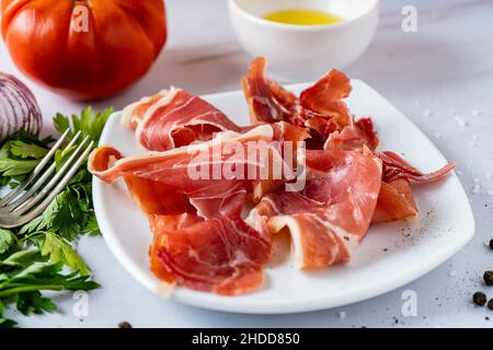 Roher oder spanischer serrano-Schinken auf einem weißen Teller auf einem Tisch mit Bio-Tomaten und Gemüse. Hohe Ansicht. Stockfoto