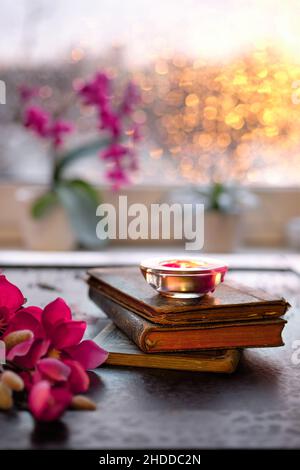 Winterzeit mit Kerze auf Stapel alter Bücher.Sonnenuntergangsfenster mit orangefarbenem Glanz, rosa und fuchsia Orchidee und Magnolienblüten. Aromatischer Teelicht Stockfoto