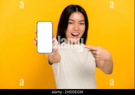 Defokussierte chinesische positive junge Frau im lässigen T-Shirt mit modernem Handy mit leerem weißen Bildschirm und zeigt einen Finger auf ihn, steht auf isoliertem gelben Hintergrund, Mockup, Kopierraum Stockfoto