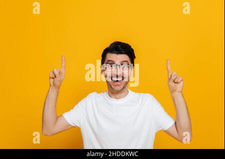 Positiver kaukasischer junger Mann in weißem Basic-T-Shirt und Brille erstaunt blickt auf die Kamera und zeigt die Finger auf leeren Raum, steht auf isoliertem orangefarbenem Hintergrund Stockfoto