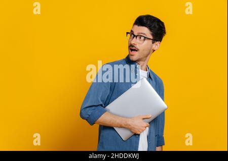 Überrascht schockiert kaukasischen stilvollen smart Kerl mit Brille, hält Laptop in der Hand, blickt mit Erstaunen in Richtung leeren Raum, auf isolierten orangefarbenen Hintergrund stehend Stockfoto