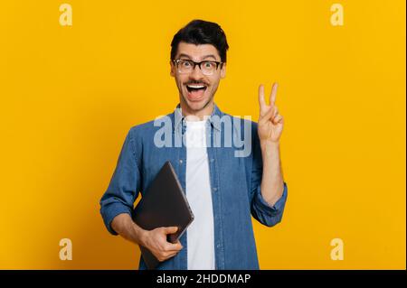 Freundlich positiv kaukasisch stilvoll schöner Kerl mit Brille, Freiberufler oder Student, hält einen Laptop in der Hand, zeigt eine friedliche Geste, steht auf isoliertem orangefarbenem Hintergrund, blickt lächelnd auf die Kamera Stockfoto