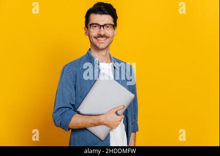 Smart schöner positiver Kaukasier, mit Brille, Student oder Freiberufler, hält einen Laptop in der Hand, steht vor isoliertem orangefarbenem Hintergrund, blickt auf die Kamera und lächelt freundlich Stockfoto