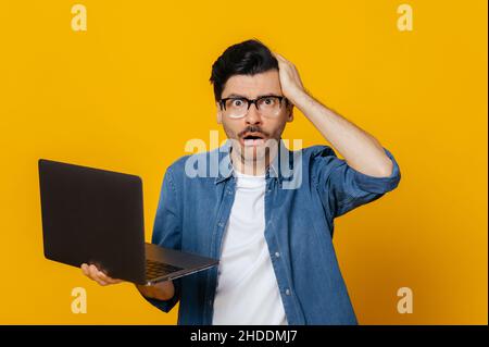 Verwirrt unzufrieden unrasiert kaukasischer Kerl in Brille und Denim-Shirt, hält einen Laptop, schaut verwirrt auf die Kamera, hält seinen Kopf, ohne Stimmung, steht auf einem isolierten orangen Hintergrund Stockfoto