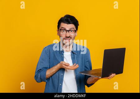 Entmutigter unrasierter Kaukasier in Brille und Jeanshemd, hält einen Laptop, schaut verwirrt auf die Kamera, hält seinen Kopf, ohne Stimmung, steht auf einem isolierten orangen Hintergrund Stockfoto
