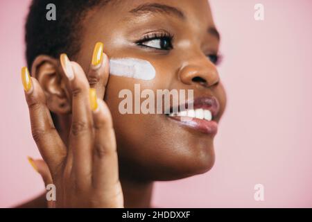 Nahaufnahme des Gesichts einer afrikanischen Frau. Anwendung Putting Creme auf Gesicht tun tägliche Routine isoliert über rosa Hintergrund. Gesichtspflege. Schönheit Stockfoto