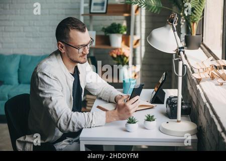 Seitenansicht eines Mannes mit Smartphone, der Notizen im Copybook aufschreibt, während er an der Universität lernt. Junger Mann mit Smartphone. Mobile Kommunikation Stockfoto