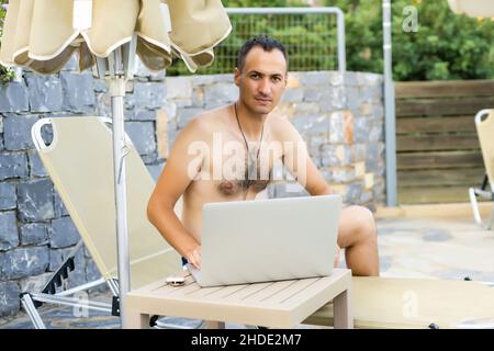 Mann mit Laptop, der sich auf der Sonnenliege am Pool entspannt Stockfoto