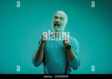Wütend frustrierter älterer Mann, der seine Fäuste ballt und seine Zähne knirscht, während er auf einen blauen Hintergrund blickt Stockfoto