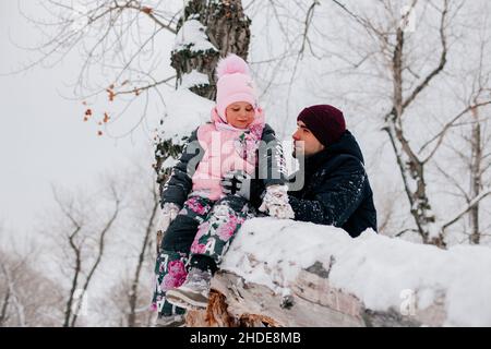 Entferntes Foto eines weiblichen Kindes, das auf Baumstamm sitzt und lächelt, in rosa Winterkleidung, während der Vater die Tochter im Wald hält. Erstaunlicher Hintergrund Stockfoto