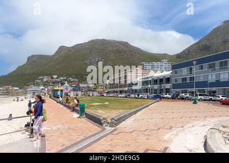 KAPSTADT, SÜDAFRIKA - 23. DEZ 2021: Eine Strandszene auf Muizenberg in Kapstadt, Western Cape Province in Südafrika. Gebäude, Fahrzeuge und Pöppeln Stockfoto