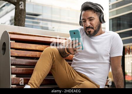 ein lateinischer Mann mit Bart sitzt auf einer Bank und blickt in einer städtischen Umgebung auf sein Handy Stockfoto