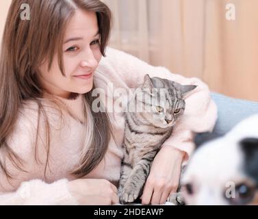 Junge Frau umarmt ihre Katze auf dem Bett zu Hause. Frau und Katze liegen auf einem Bett. Grau tabby ruhige Katze und lächelnde glückliche Dame Stockfoto
