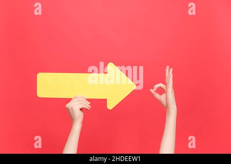 Weibliche Hände mit gelbem Pfeil zeigen auf OK-Geste, zeigen Zustimmung oder Zustimmung, isoliert über roter Wand mit Platz für Werbeaktionen Stockfoto